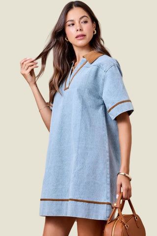 A woman modeling a light blue short-sleeve tunic dress with brown trim details, standing against a solid beige studio background and holding a brown handbag.