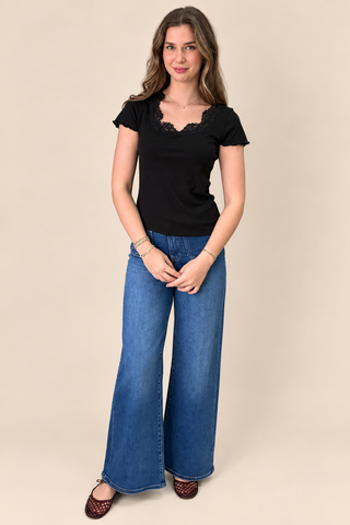 A woman modeling a black short-sleeve top with a lace-trimmed neckline and wide-leg blue jeans against a solid beige studio background.