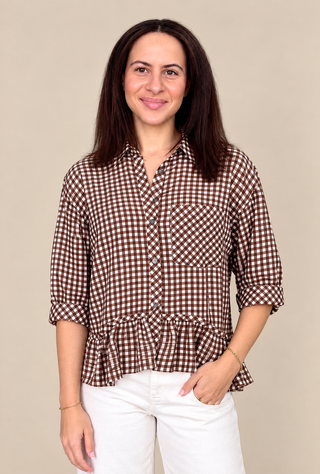 A woman modeling a brown and white gingham button-up shirt with a ruffled peplum hem, paired with white pants, against a solid beige background.
