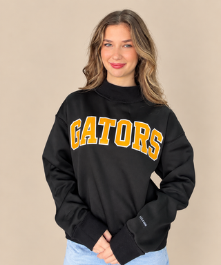 A woman wearing a black sweatshirt with 'GATORS' printed in yellow and white arched letters across the chest, paired with light blue jeans, set against a solid beige background.