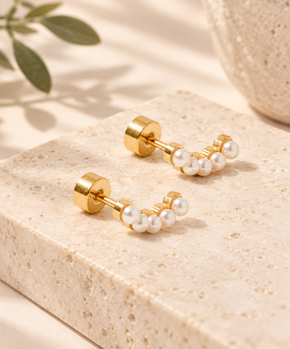 A pair of gold stud earrings with small white pearl clusters, displayed on a beige stone block with soft shadows and a blurred green leaf in the background.