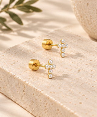 A pair of gold stud earrings with small clusters of white faux pearls, displayed on a textured beige stone block with soft shadows and a leaf in the background.
