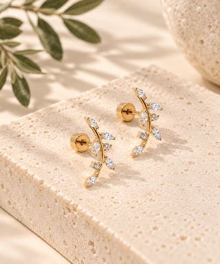A pair of gold leaf-shaped stud earrings with clear marquise and oval gemstones, displayed on a textured beige stone block with soft-focus olive leaves in the background.