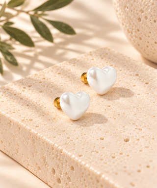 Two small heart-shaped pearl stud earrings with gold posts, displayed on a textured beige stone block with soft shadows and a leafy sprig in the background.