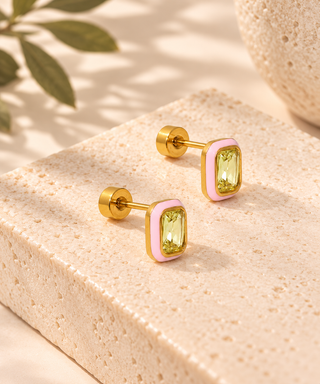 A pair of gold-toned stud earrings with rectangular light green gemstones and a soft pink enamel border, displayed on a textured beige stone block with warm natural shadows.