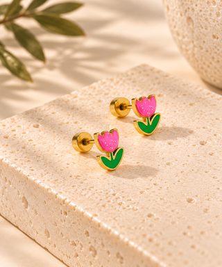 A pair of gold-tone stud earrings shaped like tulips, with pink enamel petals and green leaves, displayed on a textured beige stone block.