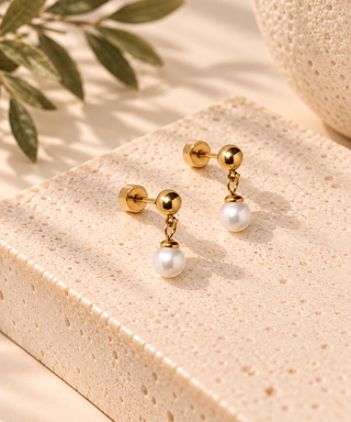 A pair of gold stud earrings with a small gold ball, a short gold chain, and a single white pearl drop, displayed on a textured beige stone block with soft shadows and green leaves in the background.