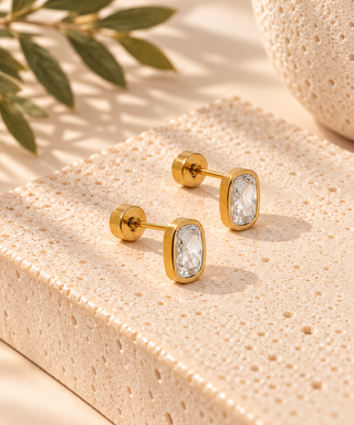 A pair of gold-toned rectangular stud earrings with clear marquise-cut stones, displayed on a textured beige stone block with soft shadows and green leaves in the background.