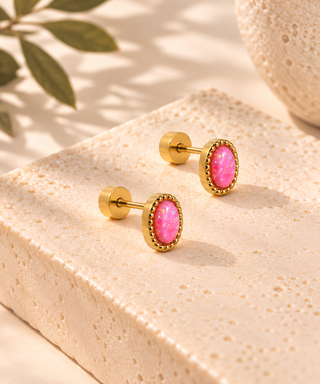 A pair of gold post earrings with oval pink cabochon stones, displayed on a textured beige stone block with soft shadows and a leaf in the background.