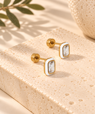 A pair of gold-toned rectangular stud earrings with clear faceted gemstones and white enamel accents, displayed on a textured beige stone block with soft shadows and a leaf in the background.