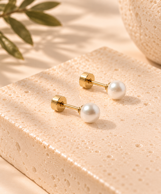 A pair of gold post earrings with round white pearl studs, displayed on a textured beige stone block with soft shadows and a leaf in the background.