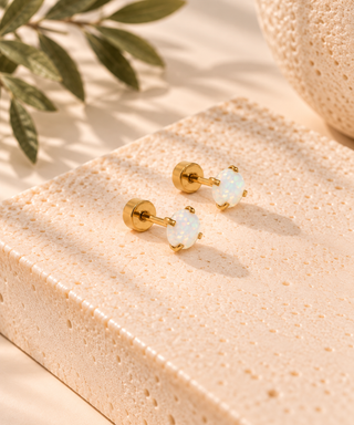 A pair of gold stud earrings with small oval white opal gemstones, displayed on a textured beige stone slab with soft shadows and decorative leaves in the background.