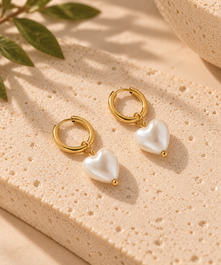 A pair of gold hoop earrings with dangling white pearl heart charms, displayed on a textured beige stone slab with soft shadows and green leaves in the background.
