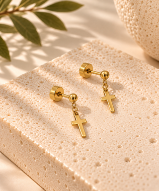 A pair of gold cross dangle earrings displayed on a textured beige stone block with soft shadows and a leaf in the background.