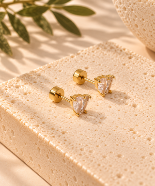 A pair of gold post earrings with pear-cut clear gemstones, displayed on a textured beige stone block with soft shadows and green leaves in the background.