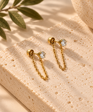 A pair of gold chain drop earrings with clear gemstones, displayed on a textured beige stone block with soft shadows and a leaf in the background.