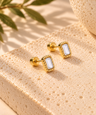 A pair of gold rectangular stud earrings with clear baguette-cut gemstones, displayed on a textured beige stone block with soft shadows and blurred green leaves in the background.