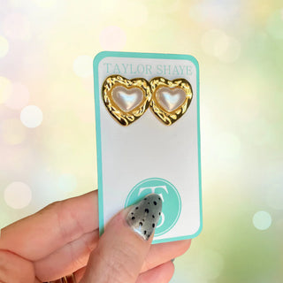 Heart-shaped earrings on a Taylor Shaye card held by a hand with black and white nail design.