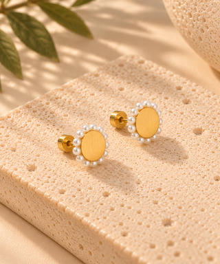 A pair of gold oval stud earrings with a border of small white pearls, displayed on a textured beige stone block with soft shadows and green leaves in the background.