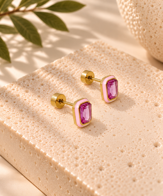 A pair of gold-toned stud earrings with rectangular pink bezel-set purple gemstones, displayed on a textured beige stone slab with soft shadows and a leaf in the background.