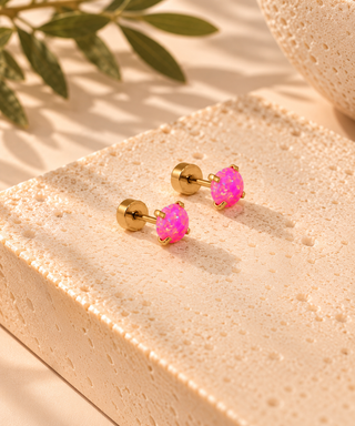 A pair of gold stud earrings with vibrant pink iridescent cabochon stones, displayed on a textured beige stone block with soft shadows and green leaves in the background.