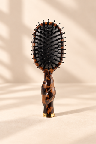 A classic oval hairbrush with a glossy tortoiseshell handle and black bristles, standing upright on a beige surface with soft leaf shadows.