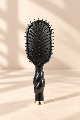A black paddle hairbrush with a sculpted twisted handle and a gold accent at the base, standing upright on a beige surface with soft shadow overlays.