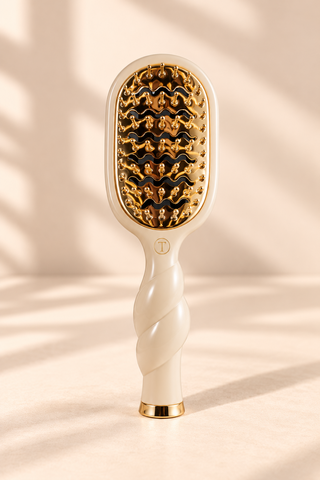 A handheld scalp massage brush with a cream-colored handle and a gold and black textured bristle head, standing upright on a beige surface with soft shadow patterns.