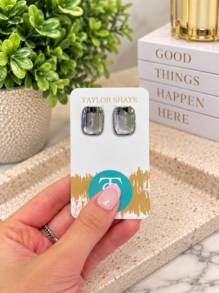 A hand holds a white jewelry display card featuring a pair of square stud earrings with iridescent purple-blue stones and silver settings. The card is branded Taylor Shaye, with a gold logo and gold brushstroke design near the bottom.