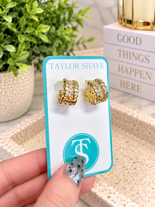 A pair of gold huggie hoop earrings with clear crystals, displayed on a turquoise and white Taylor Shaye card held by a hand. The background features a potted plant and a stack of books.