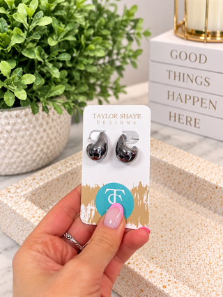 A hand with pink nail polish holds a display card featuring a pair of dangling teardrop-shaped earrings. The earrings are dark metallic with a reflective silver-toned edge.