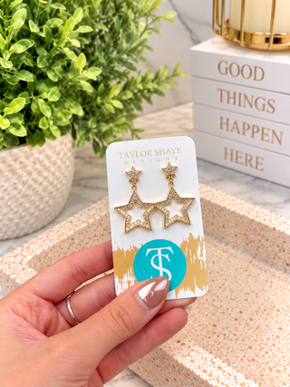 A hand holds a white jewelry display card featuring a pair of gold star-shaped dangle earrings with rhinestone trim. The card shows the brand name Taylor Shaye Designs.