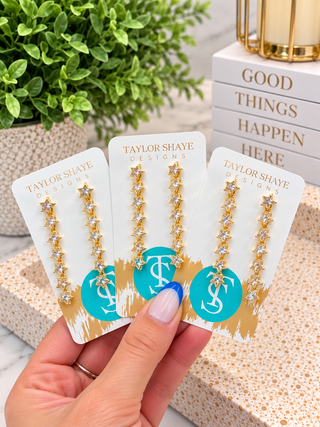 Three gold star drop earrings with clear rhinestones on white display cards branded Taylor Shaye Designs, held in a hand against a styled background with a plant and books.