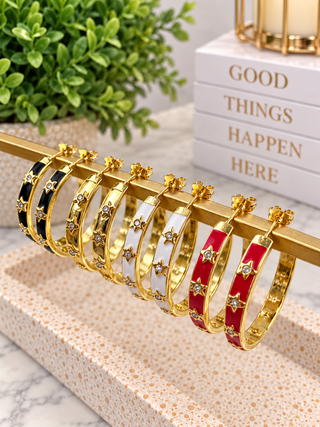 A set of gold hoop-style bangles with star and flower motifs, shown hanging on a gold display stand. The bangles have alternating black, white, and red enamel sections.