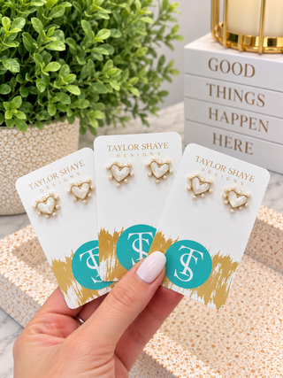 A hand holds three white jewelry display cards featuring gold heart stud earrings with pearl-like accents. The cards are branded Taylor Shaye Designs, with a potted green plant and stacked books in the background.