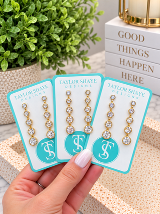 Three pairs of gold drop earrings with clear gemstones are displayed on turquoise and white cards held by a hand. The background features a potted green plant and a stack of books.