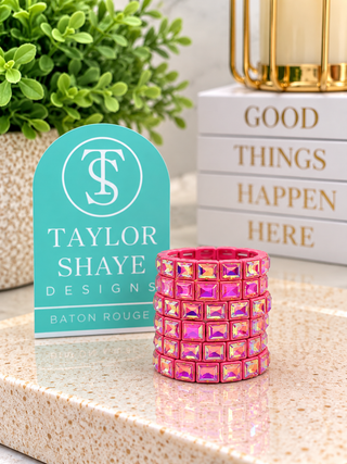 A pink rhinestone-covered wrap bracelet sits on a speckled beige countertop next to a turquoise sign reading Taylor Shaye Designs Baton Rouge, with decorative books and greenery in the background.