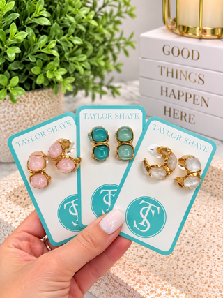 Three pairs of gold-toned statement earrings displayed on teal and white cards held by a hand. The earrings feature pink, mint green, and clear stone clusters, with the brand name 'TAYLOR SHAVE' visible on the cards.