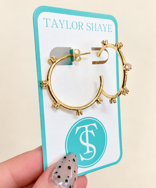 Gold hoop earrings with small crystal accents, displayed on a turquoise and white Taylor Shaye jewelry card, held by a hand with a polka-dot manicure.