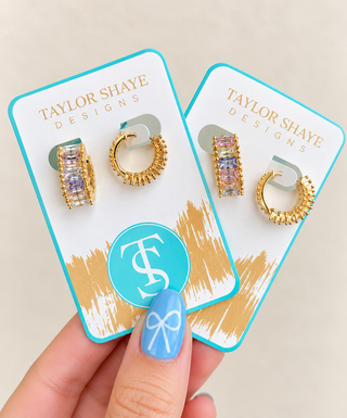 A hand with blue nail art holds two gold hoop earrings with rectangular pavé crystals on branded Taylor Shaye Designs display cards against a light beige background.