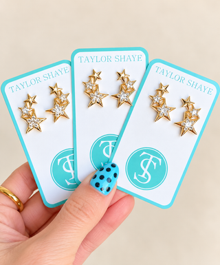 Three gold star stud earrings with clear rhinestones, displayed on white jewelry cards with turquoise borders. A hand with blue polka-dot nail polish holds the cards against a light beige background.