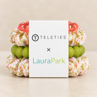 A white Teleties x LauraPark box centered in front of two fabric hair scrunchies. The scrunchies have a pink and green floral pattern with translucent coral-red and green beads.