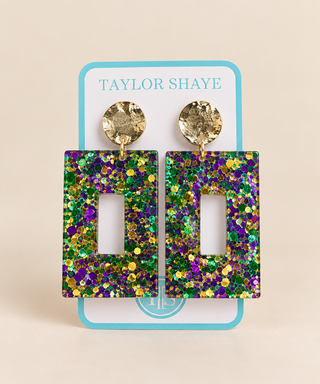 A pair of rectangular drop earrings covered in Mardi Gras glitter, hanging from textured gold studs on a light blue display card labeled 'TAYLOR SHAYE'.