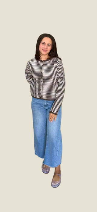 A woman stands in a clothing store aisle wearing a gray patterned long-sleeve top and light blue wide-leg jeans, surrounded by racks of clothes.