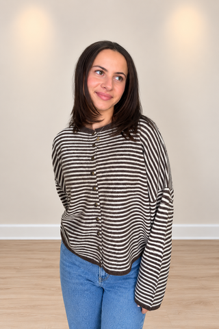 A woman with dark shoulder-length hair wearing a brown and white horizontally striped button-up cardigan and blue jeans, standing in a room with a beige wall and light wood floor.