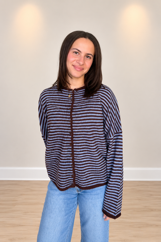 A woman wearing a navy and dark brown horizontally striped zip-up cardigan with dark brown trim, paired with light blue jeans, standing in a room with a beige wall and light wood floor.