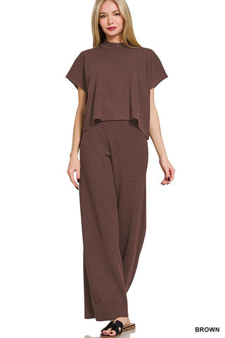 A woman modeling a matching brown two-piece set consisting of a short-sleeve mock-neck top and wide-leg pants, shown against a plain white background.