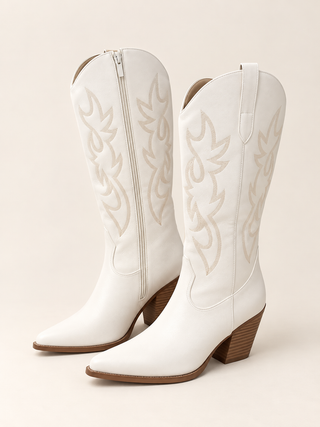 A pair of white leather cowboy boots with tan western-style embroidery and brown stacked block heels, shown against a solid light beige background.