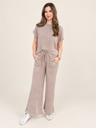 A woman modeling a matching two-piece lounge set in a textured taupe fabric, featuring a short-sleeve crewneck top and wide-leg drawstring pants, paired with white sneakers against a solid light beige background.