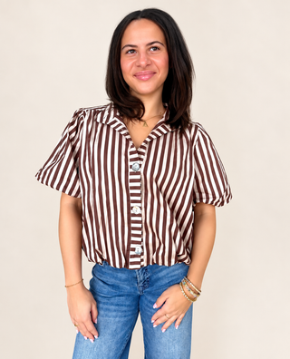A woman modeling a brown and white vertically striped short-sleeve button-up shirt with white buttons, paired with blue jeans, against a solid light beige background.